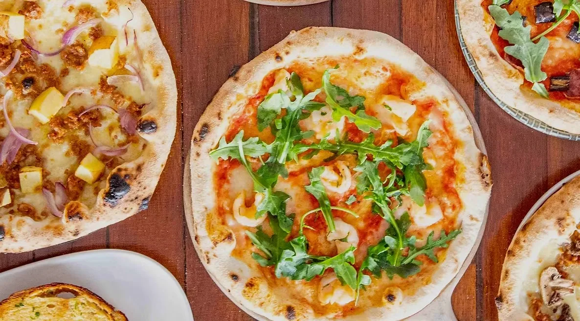 Overhead view of pizzas from Pizza by Soho on a timber table, one topped with prawns and rocket